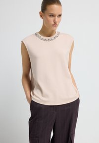 Frau in einem ärmellosen, hellrosa Oberteil mit besticktem Ausschnitt und dunklen Streifenhosen, die mit den Händen in den Taschen dasteht und zur Seite schaut.