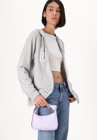 Siva zip-up kapuca s belim natpisima, uparen s visokim plavim trapericama. Mala lavanda torbica s teksturiranim drška se drži u jednoj ruci.