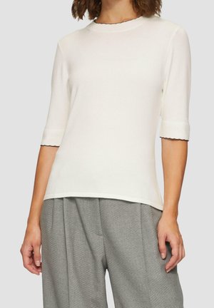 Femme portant un haut blanc à manches courtes avec un bord noir et un pantalon gris ample taille haute, posture détendue et mains le long du corps.