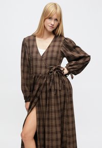Dlhé hnedé kárované zavinovacie šaty s nariasenými rukávmi, výstrihom do V a bočným rozparkom. Má zodpovedajúci opasok viazaný v páse. Mäkká tkaná tkanina.