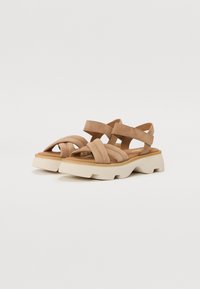 Light brown sandals feature multiple crossed straps and a cushioned footbed. The sole is chunky and off-white with textured detailing.