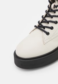 Bota de cuero blanca con punta redonda, suela de goma negra y cordones trenzados negros. Presenta detalles de costura y ojales de metal.