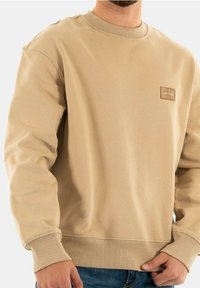 Man i beige rundhalsad sweatshirt med liten fyrkantig logotyp på bröstet, i kombination med blå jeans, mot en ljus bakgrund.