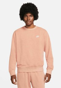 Světle růžová mikina vyrobená z měkkého materiálu. Má kulatý výstřih a malé bílé logo Nike na hrudi. Mírně textilní látka.