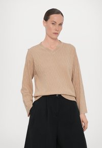 Maglione beige lavorato a maglia con scollatura a V, caratterizzato da un motivo a rombi e maniche lunghe, abbinato a pantaloni neri a vita alta.