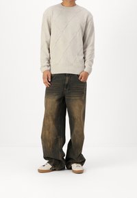 Personne portant un pull beige texturé, un jean marron délavé ample, et des baskets blanches à rayures noires, debout devant un fond blanc.