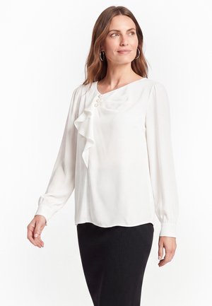 Femme portant un chemisier blanc avec des volants et des boutons en perle, et une jupe noire, regardant légèrement sur le côté sur un fond blanc.