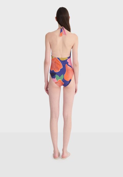 Femme portant un maillot de bain une pièce floral coloré avec un lien de cou noué, debout pieds nus sur un fond clair uni, vue de dos.