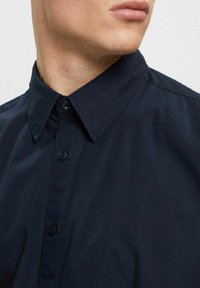 Man in een donkerblauwe button-up shirt met een klassieke kraag, zichtbaar van de nek tot halverwege de borst, hoofd iets naar de zijkant gedraaid.