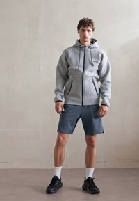 Sivi dukserica sa zatvaračem i dva prednja džepa, u paru s tamnim šortsevima i crnim sportskim cipelama. Model stoji uz zid svijetle boje.