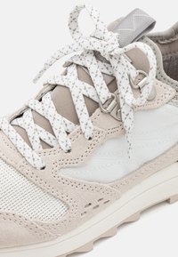 Gros plan sur une basket beige et blanche avec des panneaux en mesh et en daim, des lacets blancs à motifs noués en nœud, et une languette en tissu gris avec un passant.