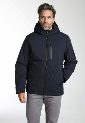 Homme portant une veste d'hiver noire à capuche avec fermeture éclair à l'avant et pantalon gris, debout devant un fond clair uni.