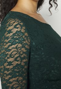 Vêtement en dentelle verte avec de longues manches transparentes, présentant des motifs floraux complexes et un corsage ajusté texturé. Détail du col lisse.