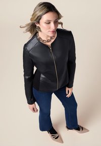 Sort læderjakke med lynlås, ribbet tekstur på ærmerne, båret over tætsiddende denim, parret med spidse beige sko og et guld kædehalssmykke.
