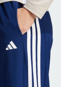 Navy athletic pants with white three-stripe design along the side. Lightweight fabric, featuring a side pocket and an Adidas logo.