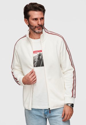 Mann trägt weiße Zip-up-Jacke mit rotem Streifen und Text auf den Ärmeln, grafisches T-Shirt und hellblaue Jeans, posiert vor grauem Hintergrund.