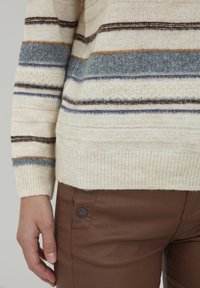 Cremfarbener gestreifter Pullover mit blauen, braunen und orangefarbenen Akzenten; strukturierter Strick. Braune lederähnliche Hosen mit seitlichem Knopfdetails.