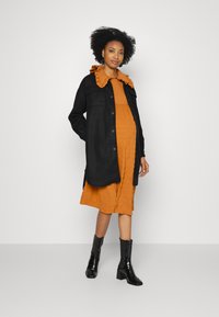 Casaco preto de botão com grandes bolsos por cima de um vestido texturizado na cor ferrugem, apresentando um colarinho com babados, combinado com botas de tornozelo pretas e brilhantes.