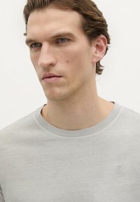 Lichtgrijze crewneck t-shirt van zachte stof, met een klein paarse schedel logo op de linkerborst, gladde textuur, eenvoudig ontwerp.