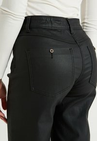 Primer plano de una persona que lleva pantalones negros con bolsillos traseros, uno de los cuales tiene un botón, y una camisa blanca de manga larga metida por dentro.