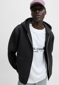 Sort zip-up hoodie med en stor hætte, frontlommer og glat tekstur, lagdelt over en hvid skjorte med trykt tekst.