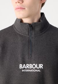 Tmavě šedá mikina na zip vyrobená z jemné látky, s vysokým límcem a bílým logem "Barbour International" na hrudi.