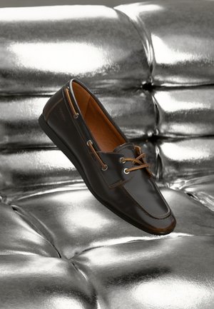 Brune skinnloafer med lysbrune aksenter og metalliske snøringer, med rund tå og glatt tekstur, plassert mot en skinnende sølvbakgrunn.