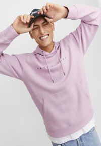 Sudadera rosa claro con un bolsillo frontal, capucha con cordón ajustable y texto en negro impreso en el pecho, combinada con jeans azul claro.