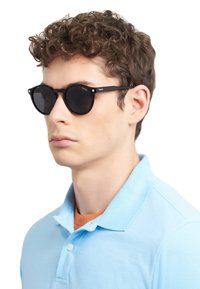 Occhiali da sole neri in stile retrò con lenti tonde e hardware minimale, indossati da una persona con capelli corti e ricci. La camicia è di colore azzurro chiaro.