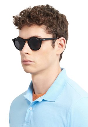 Occhiali da sole neri in stile retrò con lenti tonde e hardware minimale, indossati da una persona con capelli corti e ricci. La camicia è di colore azzurro chiaro.