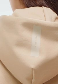 Beige hoodie met een gladde textuur. Beschikt over een rechthoekig logo-tag in het wit op de achterkant, centraal geplaatst.