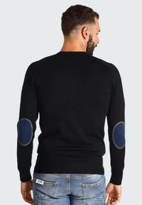 Pull noir à manches longues, texture côtelée et patchs coudes bleus. Design à encolure ronde avec une silhouette ajustée. Porté avec un jean bleu clair.