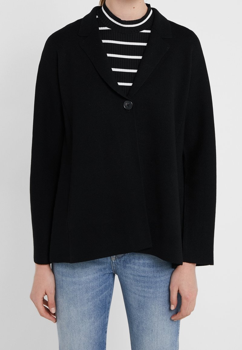 Personne portant un blazer noir à un bouton sur une chemise rayée noir et blanc avec un jean bleu clair sur un fond uni.
