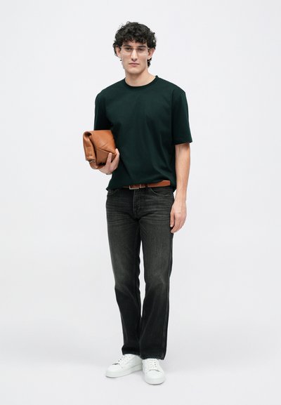 Tricou negru, blugi închiși, adidași albi. Ține o geantă din piele maro. Prezintă o tăietură lejeră și un design minimalist.