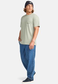 Lys grøn bomulds t-shirt med en brystlomme og blå broderi, parret med løse blå jeans og sorte sneakers, toppet med en marineblå hue.