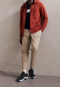 Red zip-up hoodie, black Nike t-shirt, beige tapered pants, and dark green sneakers with yellow accents. Smooth fabric textures throughout.