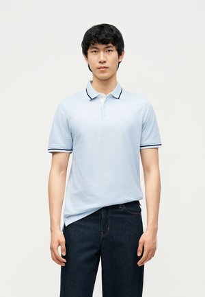 Fiatal férfi világoskék rövid ujjú galléros pólót visel sötét szegéllyel és sötétkék farmert, sima világos háttér előtt áll.