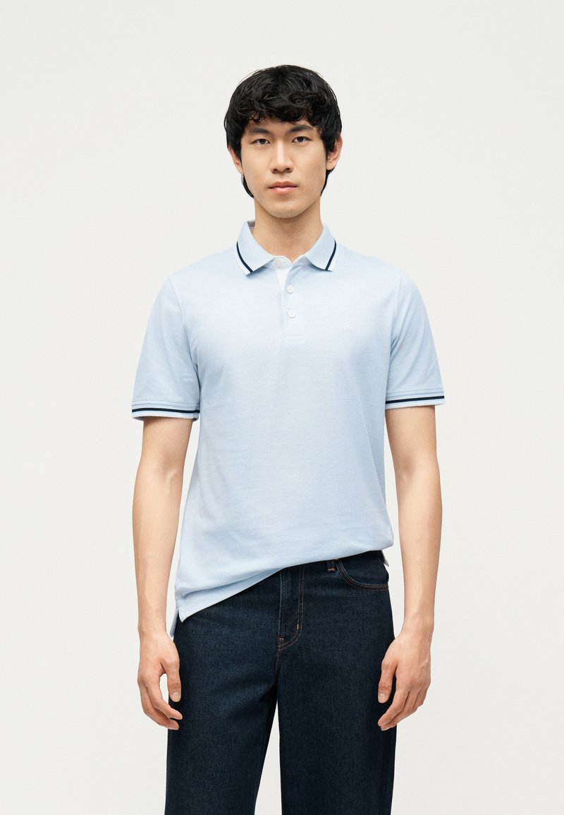 Νέος άνδρας που φοράει ανοιχτό μπλε κοντομάνικο πόλο με σκούρες λεπτομέρειες και σκούρο μπλε τζιν, στέκεται μπροστά σε απλό ανοιχτό φόντο.