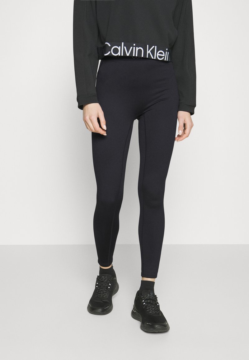 Leggings neri con un design aderente, texture liscia, alto elastico con logo Calvin Klein, abbinati a scarpe da ginnastica nere.