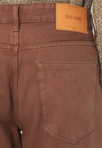 Pantaloni in denim di cotone marrone con una tasca posteriore e un'etichetta beige con la dicitura "ONLY & SONS". Il tessuto presenta una sottile trama e cuciture.