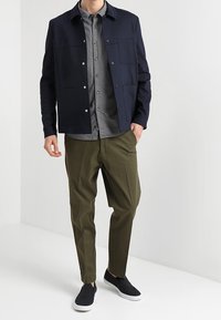 Marine prestatiesjas met drukknoopjes, grijze overhemd met knopen, olijfgroene taps toelopende broek en zwarte instap schoenen. Minimalistisch ontwerp.
