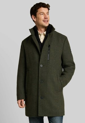 Uomo che indossa un cappotto verde scuro con bottoni e tasca sul petto con cerniera, camicia beige e jeans blu, guarda di lato con la mano in tasca.