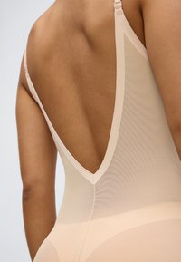 Nude body z głębokim dekoltem w kształcie litery V na plecach, wykonany z przezroczystej, teksturowanej tkaniny. Posiada regulowane ramiączka oraz dopasowany fason.