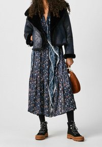 Granatowa kurtka shearling z czarnymi wykończeniami, sukienka w kwiatowy wzór, czarne botki za kostkę z masywną podeszwą oraz brązowa okrągła torebka.