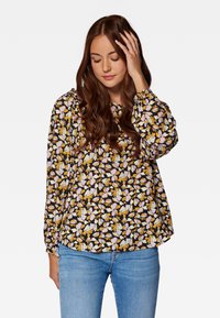 Blumenshirt in Schwarz mit lila, gelben und weißen Mustern. Verfügt über lange Puffärmel und einen runden Ausschnitt. Getragen mit hellblauen Jeans.