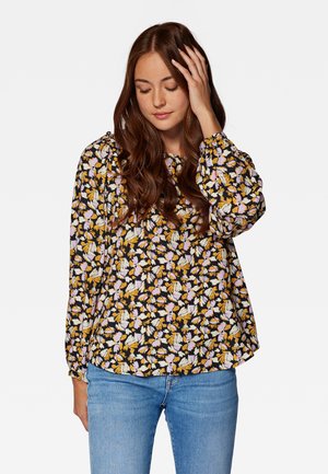 Blumenshirt in Schwarz mit lila, gelben und weißen Mustern. Verfügt über lange Puffärmel und einen runden Ausschnitt. Getragen mit hellblauen Jeans.