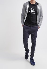 Grå zip-up hoodie över en svart T-shirt med en vit triangulär logotyp, parat med mörklila byxor och marinblå sneakers.