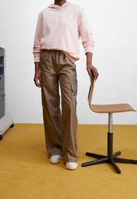 Ljusrosa huvtröja med framlogga, avslappnad passform. Ljusbruna cargobyxor med sidfickor, kombinerade med vita sneakers. Gul bakgrund på golvet.