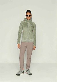 Naine, kellel on seljas helesinine kapuutsiga fliisjakker, taupe värvi püksid, hallid matkasaapad ja valged prillid, seisab ühtlase taustaga.