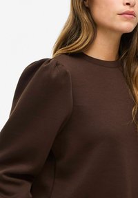 Brun genser med rund hals og puffede lange ermer, laget av et glatt, litt teksturert stoff. Ingen synlige mønstre eller accenter.